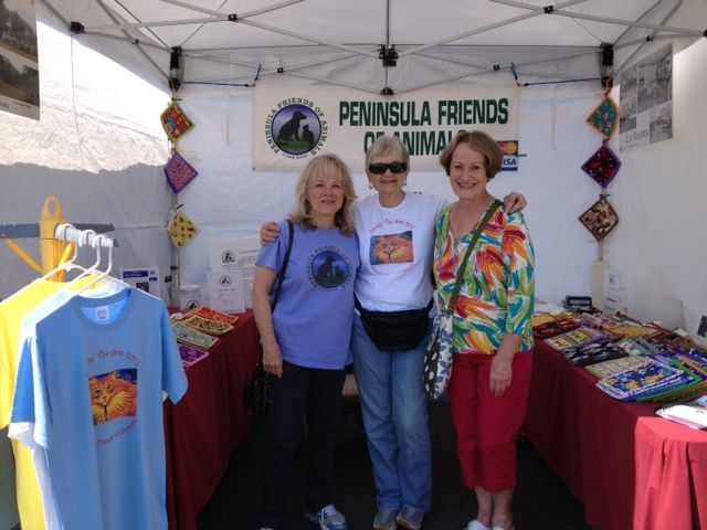 Three volunteers standing together under a white canopy at a Peninsula Friends of Animals outreach booth with displayed merchandise and a banner overhead